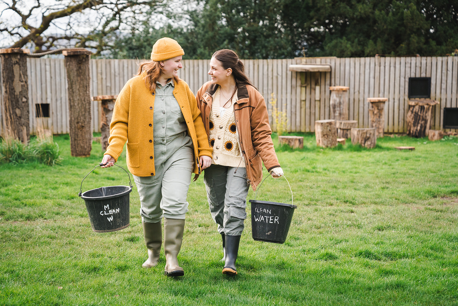 female couple walking on grass holding buckets and smiling at each other IMAGE: Rachel Hein 2024