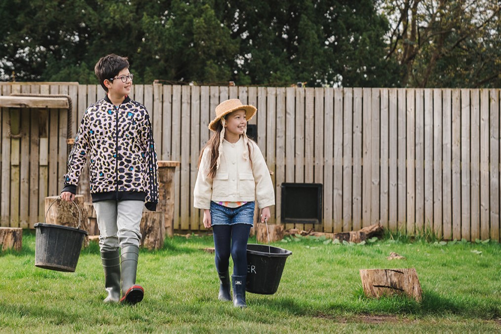 two children walking holding buckets and smiling to the side IMAGE: Rachel Hein 2024