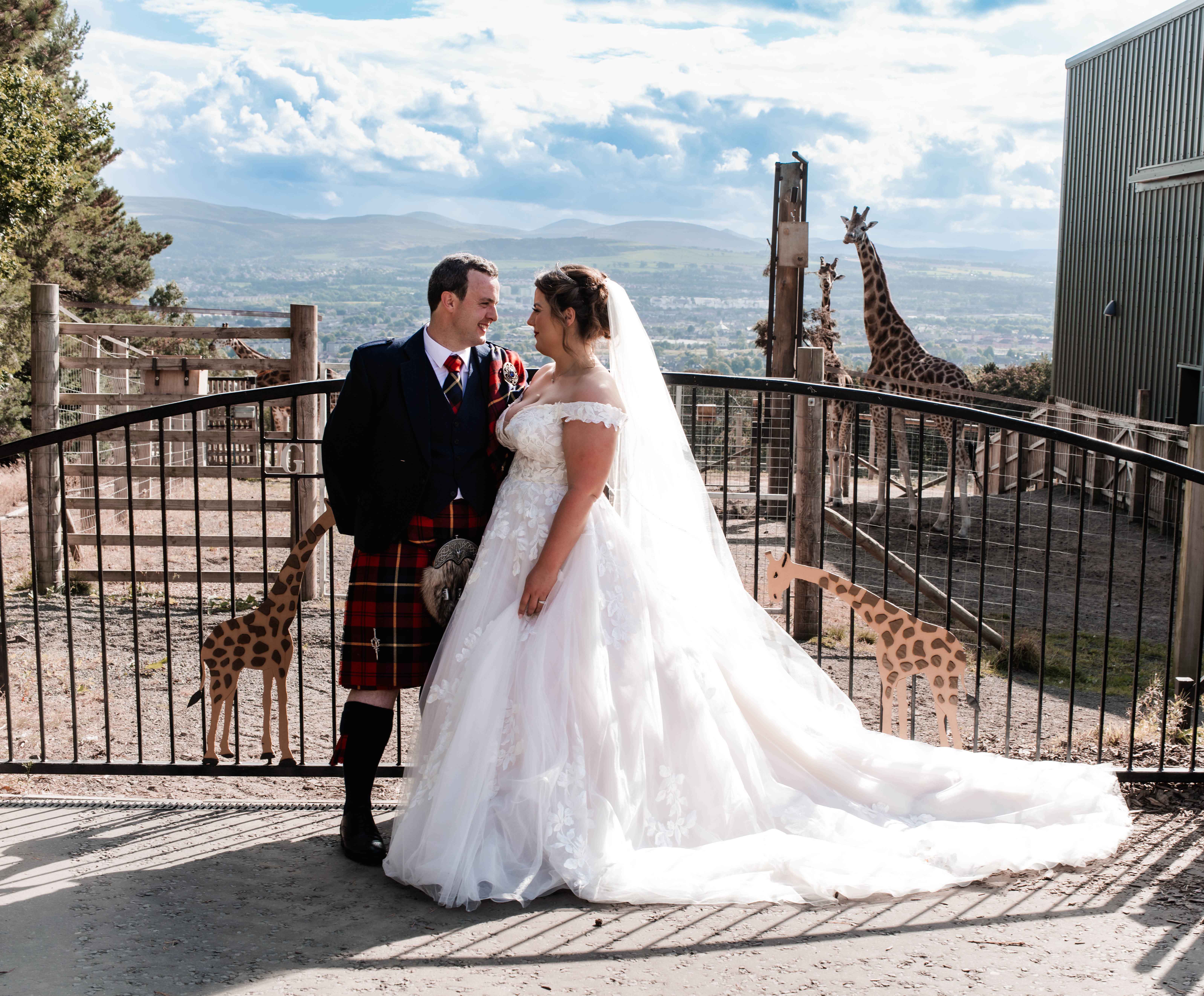 Bride and groom pose outside of giraffe gate at the side of giraffe enclosure IMAGE: Compass 2023