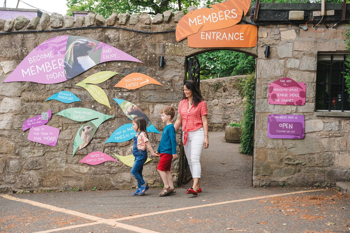 A mother and two children - a brother and sister - leave Edinburgh Zoo with smiling faces. They are exiting via the the Members Gate, which has a stone wall decorated with bright and colourful signs CREDIT RZSS 