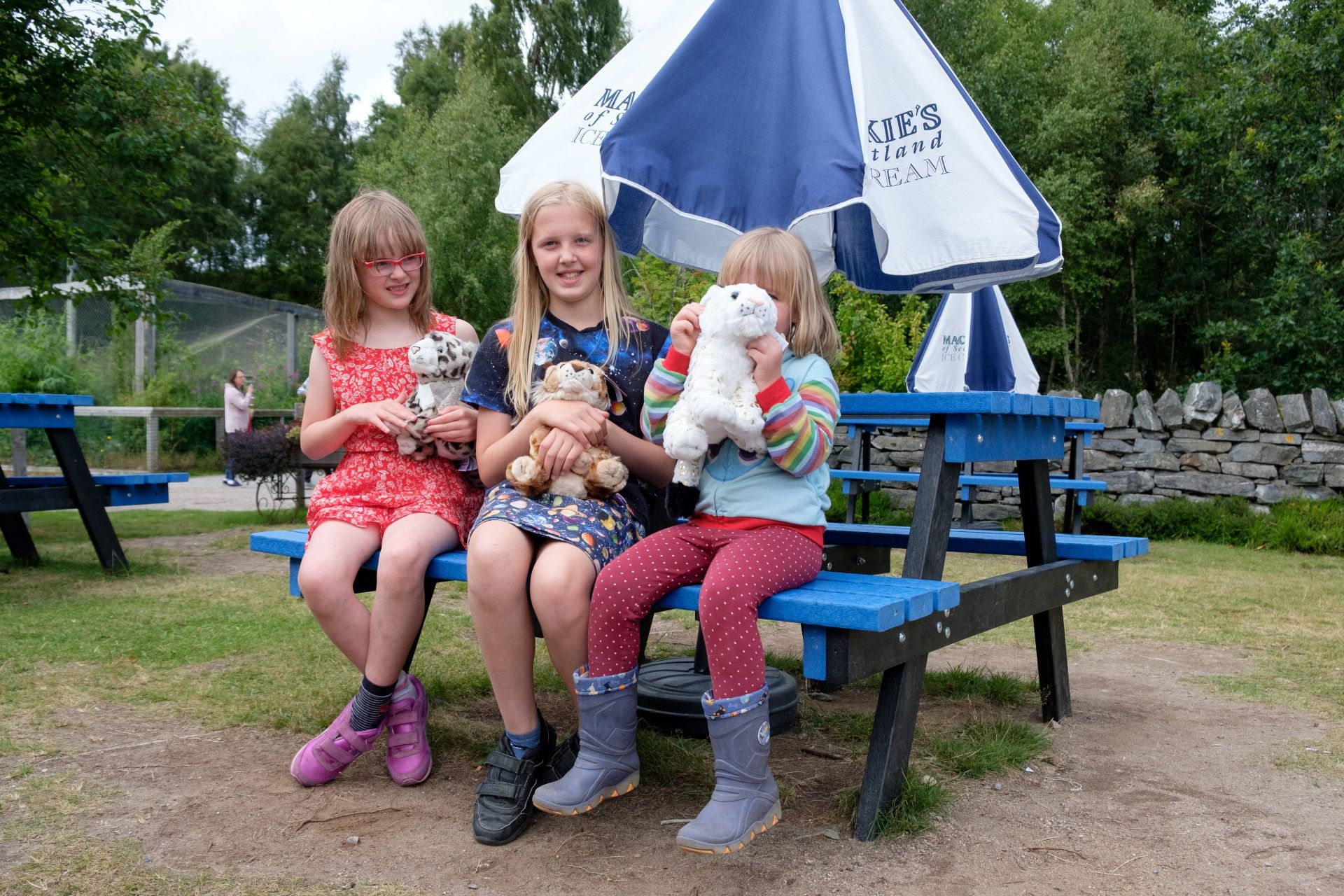 Three kids (girls) sitting on picnic bench holding soft toys from gift shop IMAGE: Robin Mair 2022